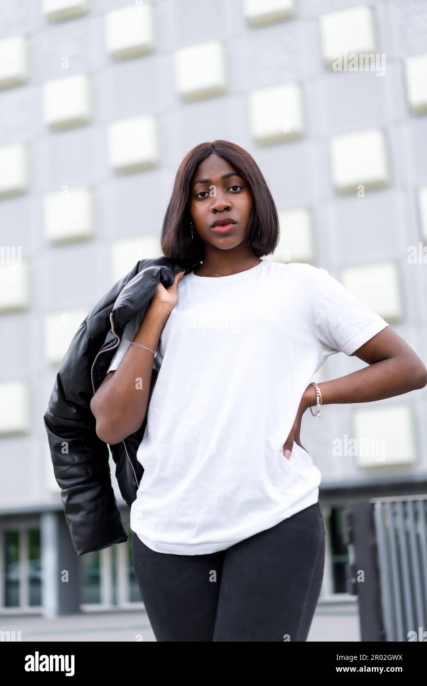 City Fashionista: Eine junge schwarze Frau posiert in einem modischen weißen T-Shirt und einer schwarzen Hose, mit einem modernen grauen Gebäude als schickem urbanen Hintergrund Stockfoto