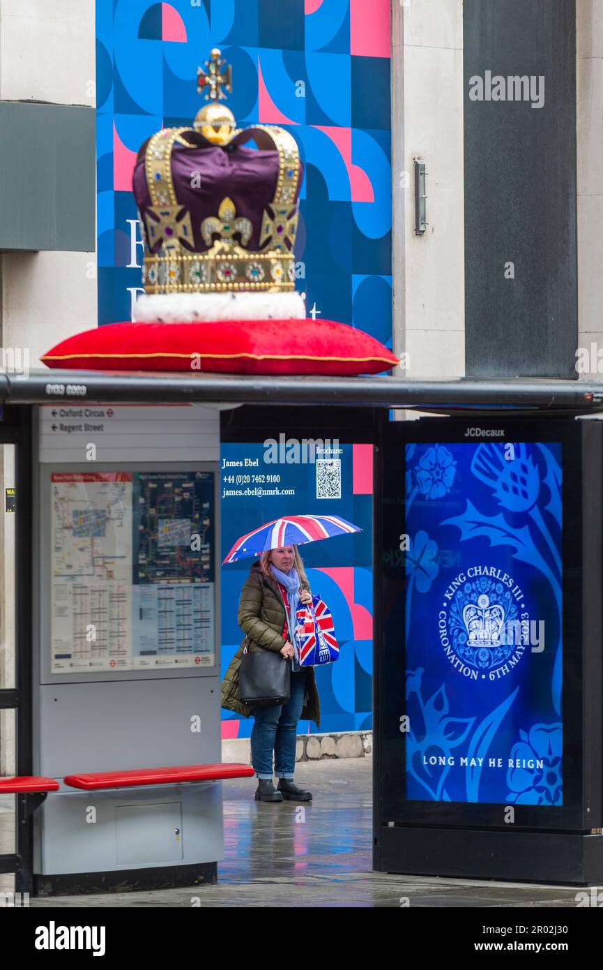 London, Großbritannien. 6. Mai 2023 Ein Tourist mit einem Union Jack-Regenschirm blickt auf eine große Krone auf dem Dach einer Bushaltestelle in der Nähe des Flagship John Lewis Kaufhauses in der Oxford Street am Tag der Krönung von König Karl III. Und Königin Camilla. Während eine digitale Nachricht auf einem Bildschirm am Busschutz angezeigt wird. Dies ist die erste Krönung des Vereinigten Königreichs seit 70 Jahren und die erste im digitalen Zeitalter. Kredit: Stephen Chung / Alamy Live News Stockfoto