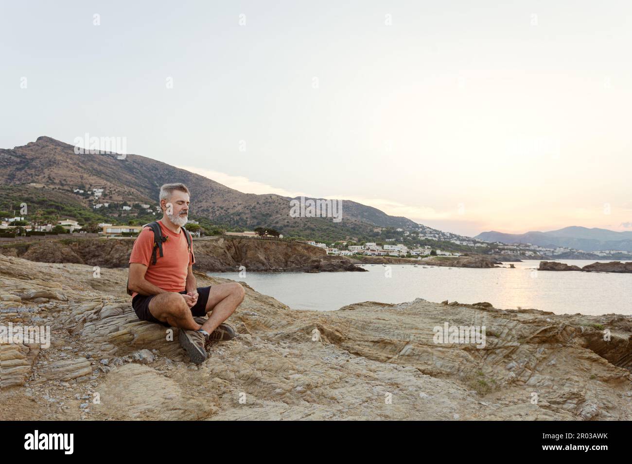 Ein erwachsener Mann mit weißem Bart, der mit geschlossenen Augen am Meer meditiert. Stockfoto