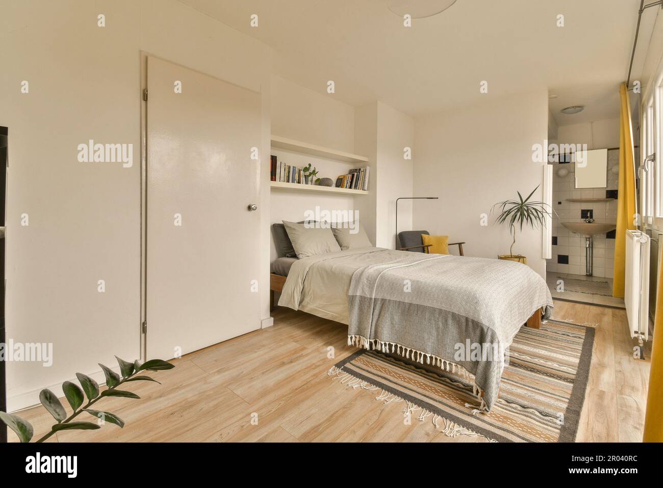 Ein Schlafzimmer mit weißen Wänden und Holzfußboden. Das Zimmer hat ein Bett und ist von gelben Vorhängen umgeben Stockfoto