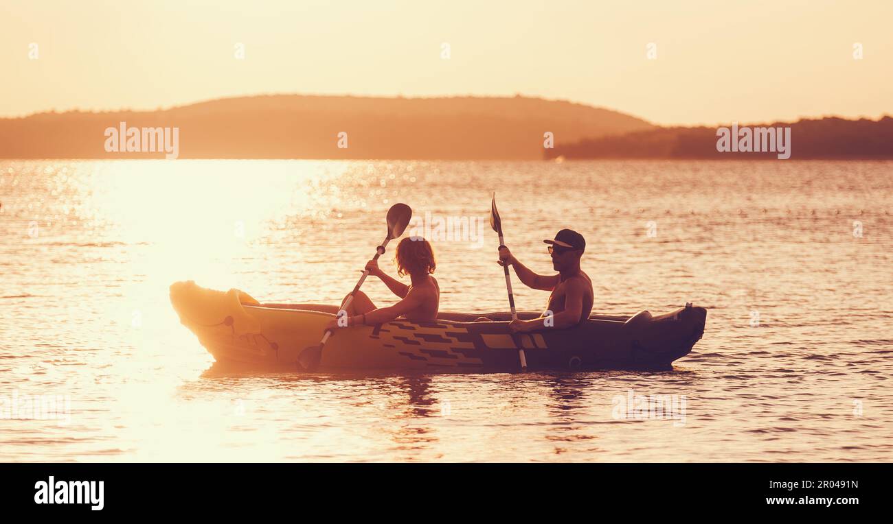 Zwei Ruderer auf einem aufblasbaren Kajak, die am Abend durch die Sonnenuntergänge rudern, strahlen den Hafen des Adriatischen Meeres in Kroatien in der Nähe von Sibenik. Urlaub, Sport und Erholung Stockfoto