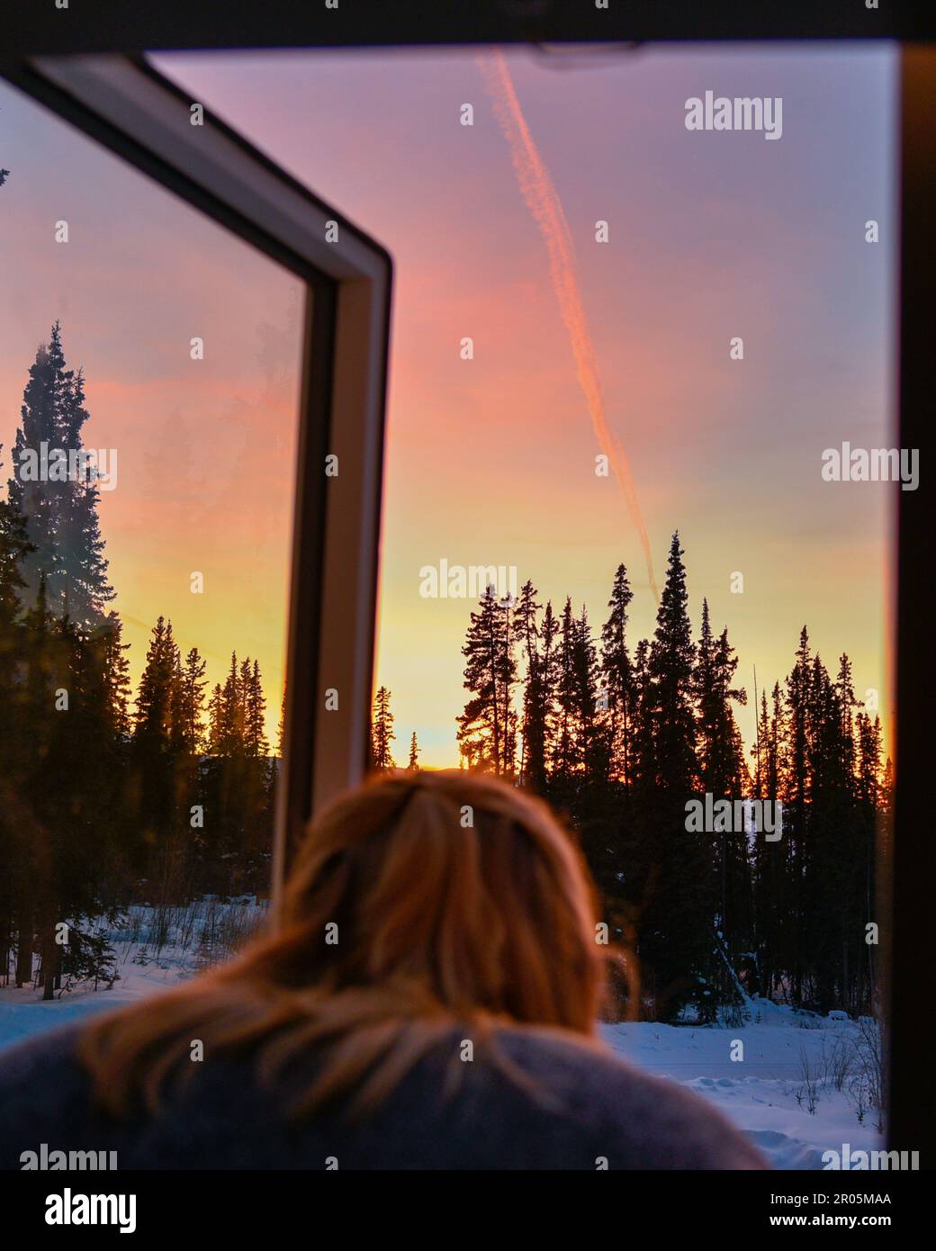 Frau, die im Herbst vor dem wunderschönen pastellrosa Sonnenuntergang steht, während sie aus einem Fenster blickt und die Schönheit der Erde bewundert. Stockfoto