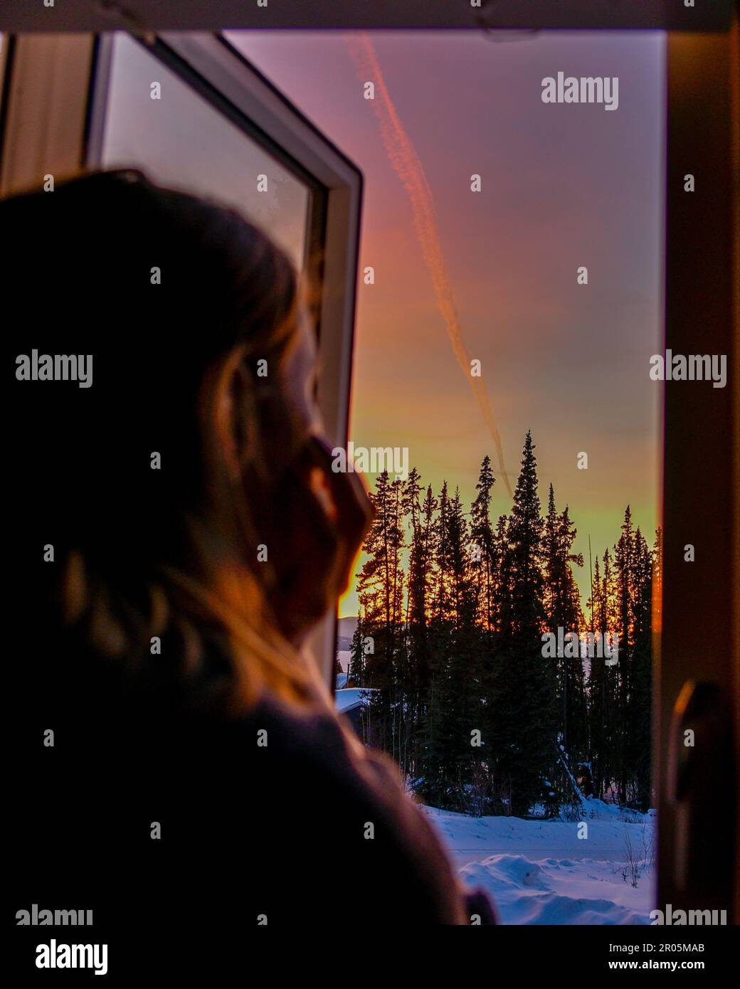 Frau, die im Herbst vor dem wunderschönen pastellrosa Sonnenuntergang steht, während sie aus einem Fenster blickt und die Schönheit der Erde bewundert. Stockfoto