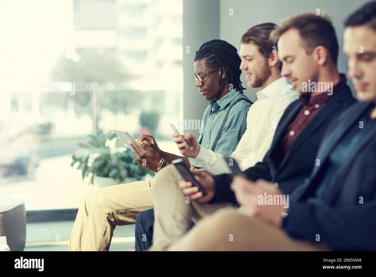 Testen ihrer Social-Networking-Fähigkeiten. Eine Gruppe von Geschäftsleuten, die verschiedene drahtlose Geräte nutzen, während sie in der Schlange auf ein Interview warten. Stockfoto