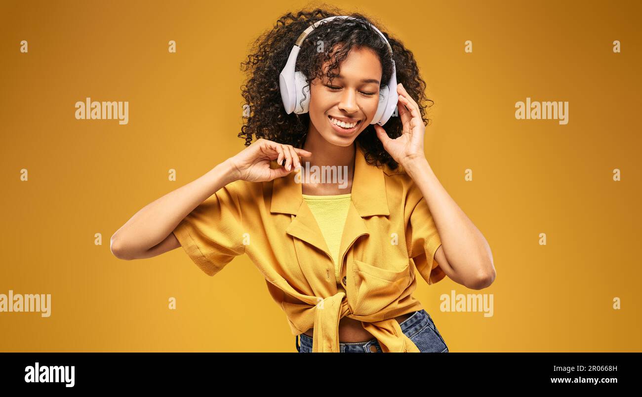 Eine afroamerikanische Frau hört gerne Musik über ihre weißen Kopfhörer, die im Sommer-Trend-Stil auf gelbem Hintergrund posieren Stockfoto
