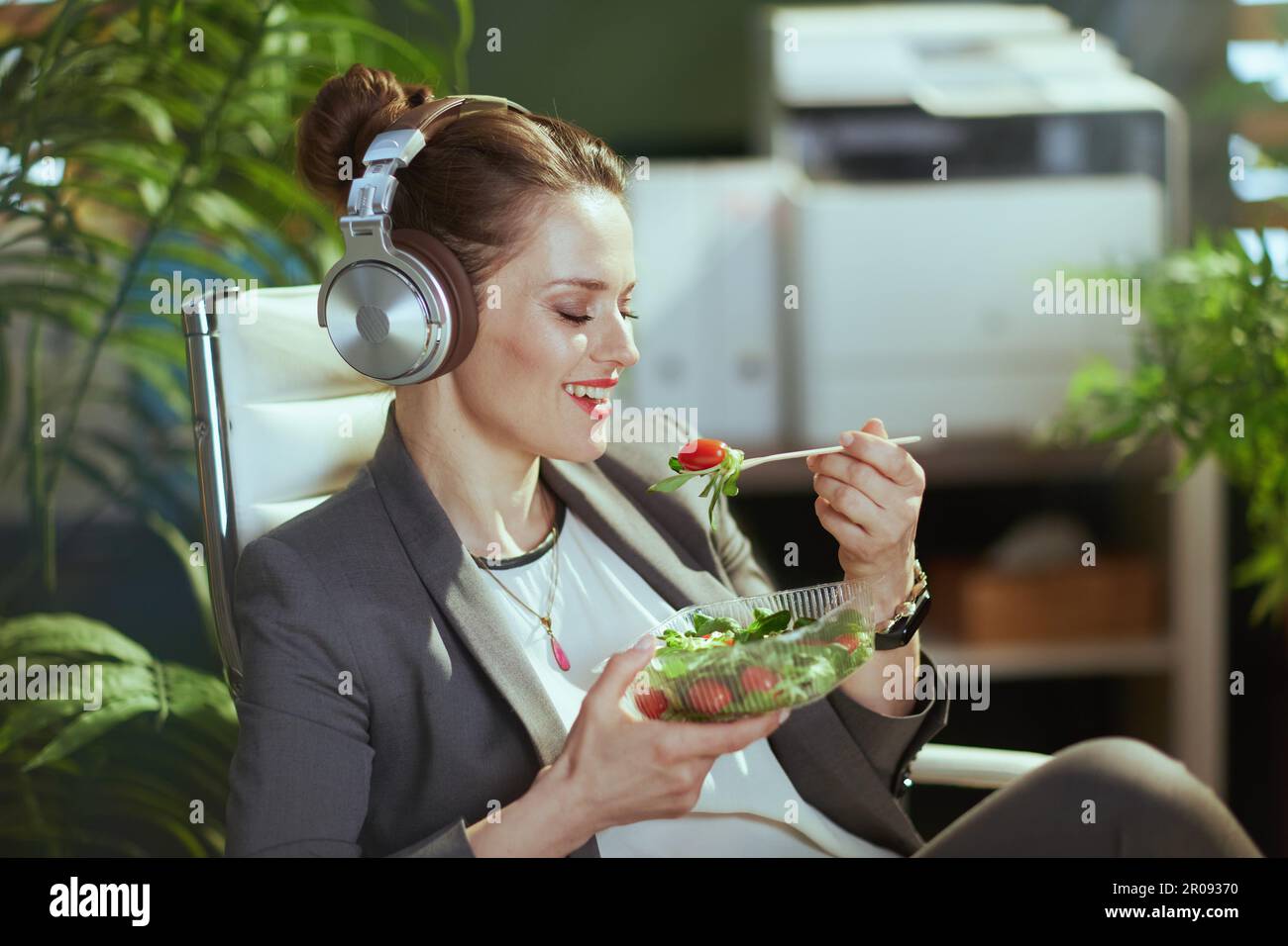 Nachhaltiger Arbeitsplatz. Lächelnde, moderne Eigentümerin eines kleinen Unternehmens in einem grauen Business-Anzug in einem modernen grünen Büro mit Kopfhörern, die Salat essen. Stockfoto