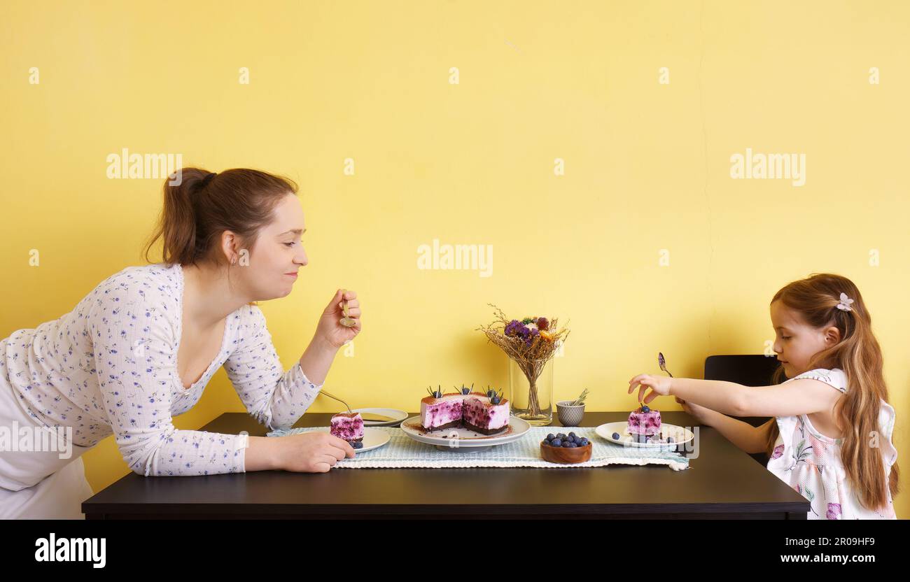 Eine junge Mutter und ihre kleine Tochter sitzen zusammen am Esstisch, essen Blaubeerkäsekuchen und genießen eine Geburtstagsparty. Stockfoto