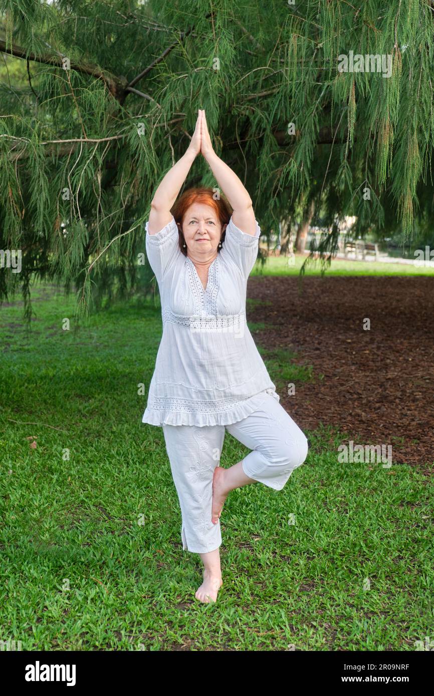 Reife hispanische Frau in weißer Kleidung, die im Park Yoga praktiziert. Vrksasana steht. Konzepte: Wellness, Vitalität, aktiv und gesund l Stockfoto