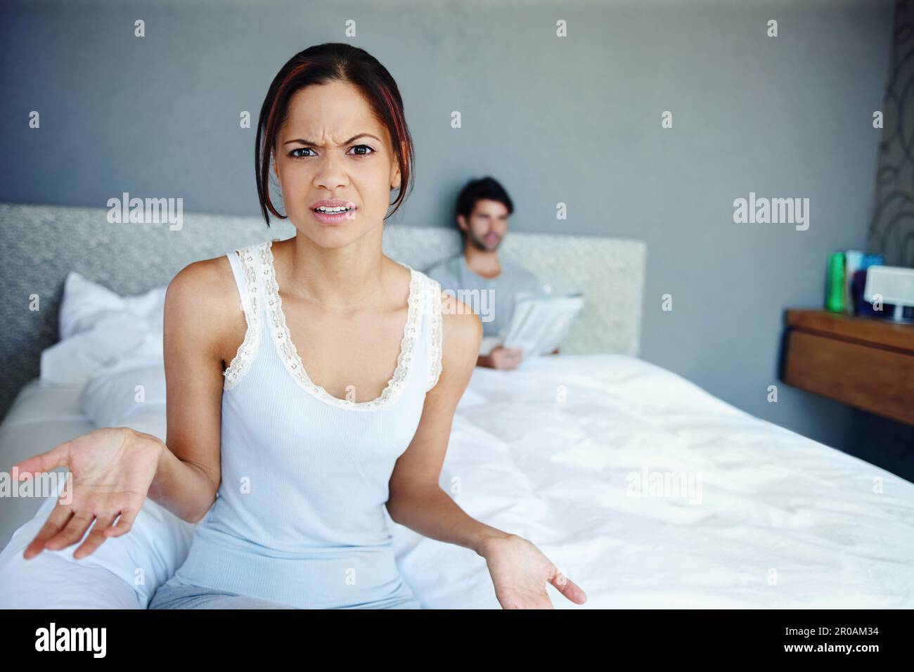 Ich bin über diese Situation verwirrt. Porträt einer verwirrten Frau, die auf der Seite ihres Bettes sitzt, mit ihrem Freund im Hintergrund. Stockfoto