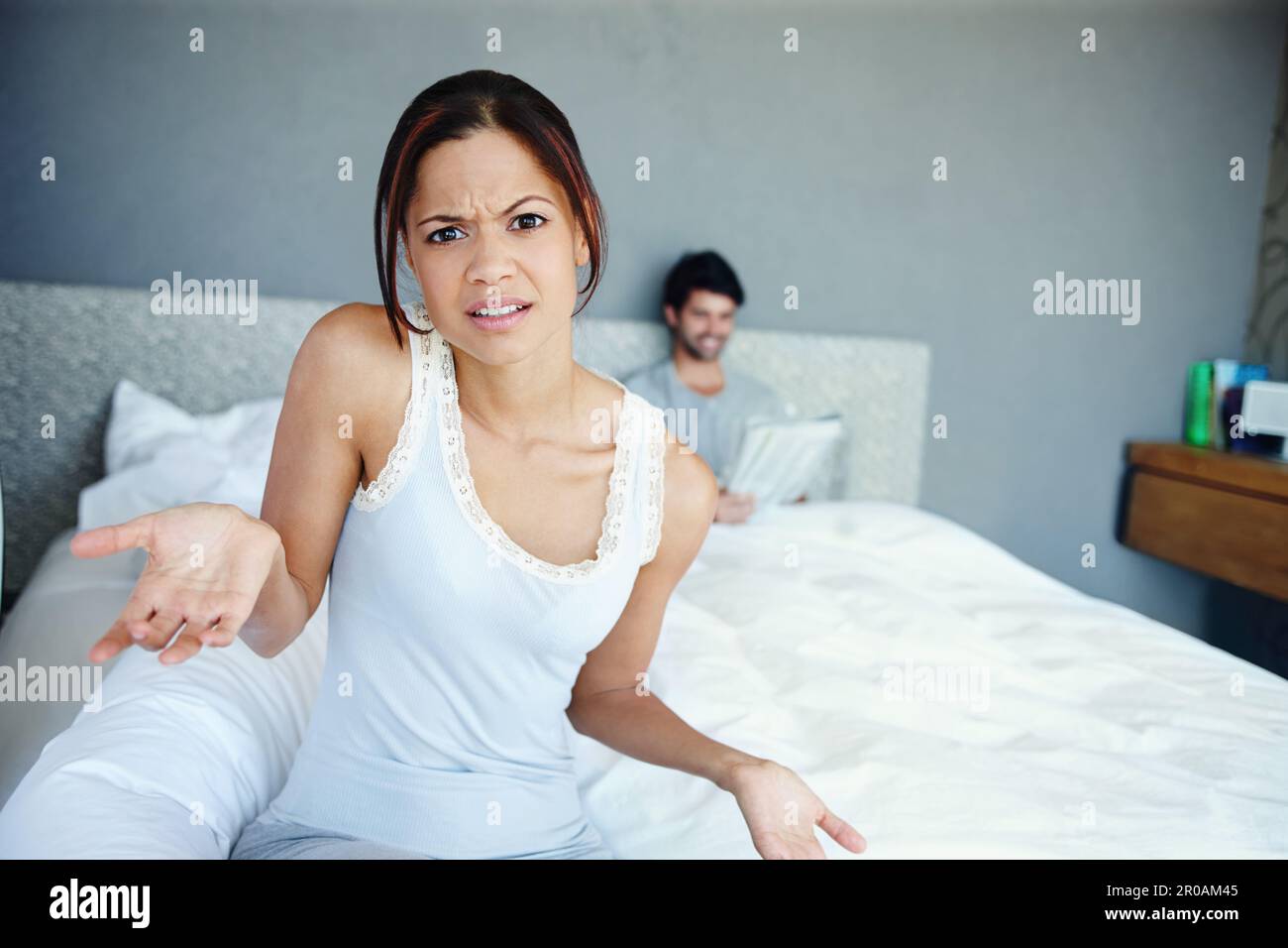 Was mache ich jetzt? Porträt einer verwirrten Frau, die auf der Seite ihres Bettes sitzt, mit ihrem Freund im Hintergrund. Stockfoto