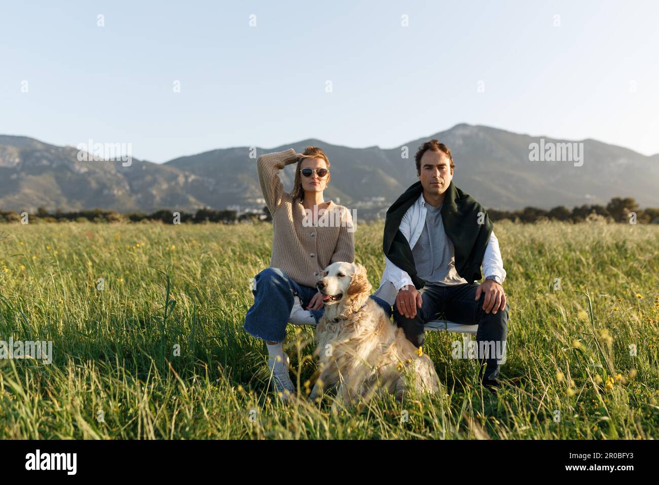 Ein junges glückliches Paar mit Hund, das auf Stühlen im Federrasenfeld sitzt, am Tag des Sonnenuntergangs im Sommer, und sich in der Kamera nach vorn freut, ernsthaft. Stockfoto