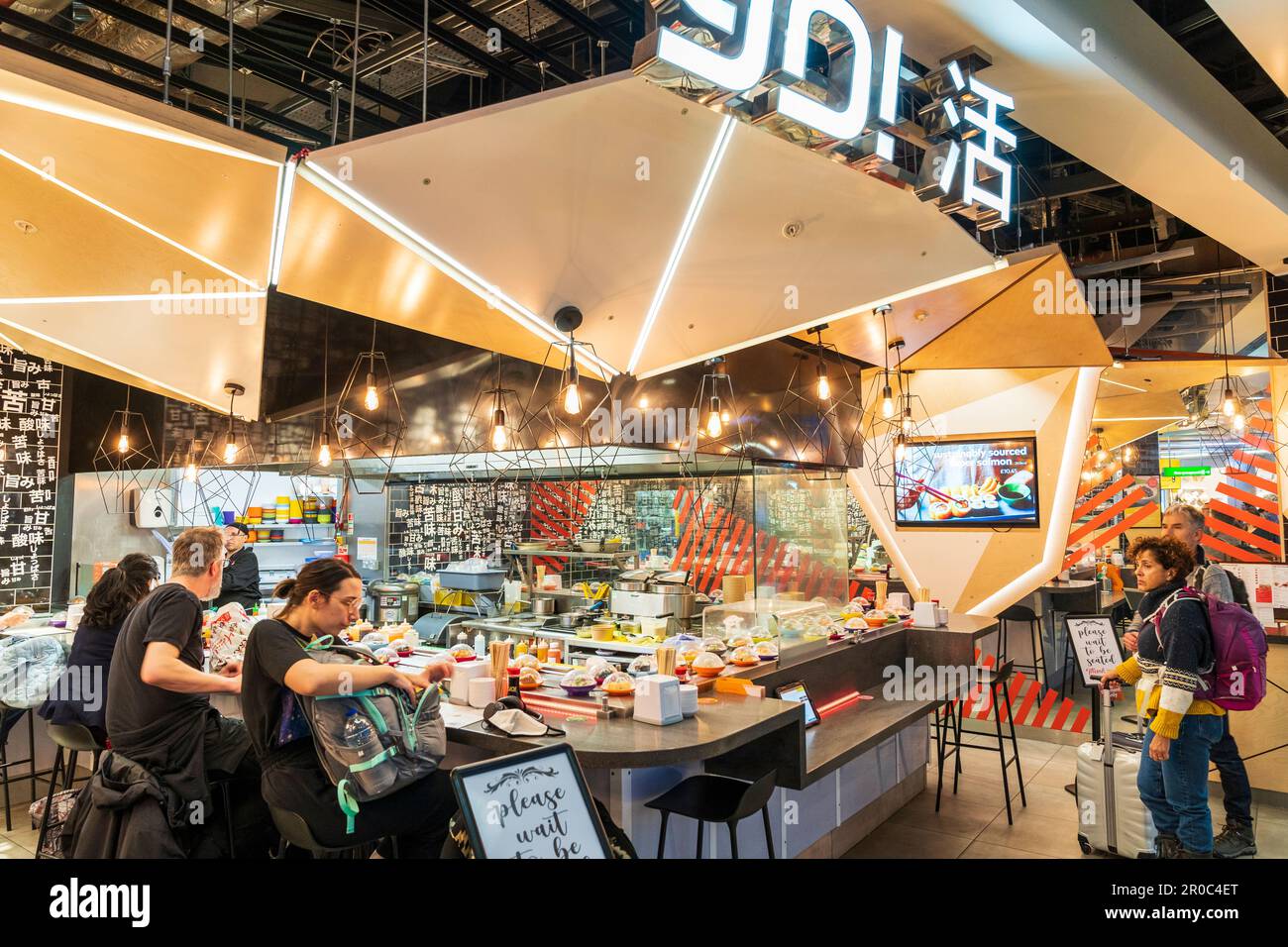 Hey! Sushi-Restaurant in der Abflughalle von Terminal 3 des Flughafens Heathrow. Zwei Leute warten auf einen Sitzplatz, andere essen an der Theke. Stockfoto