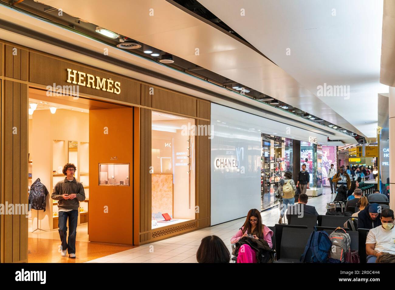 Hermes und Chanel werden in der Abflughalle des Terminals 3 am Flughafen London Heathrow eingekauft. Ein Mann verlasst den Hermes Laden Leute sitzen drauBen Stockfotografie Alamy