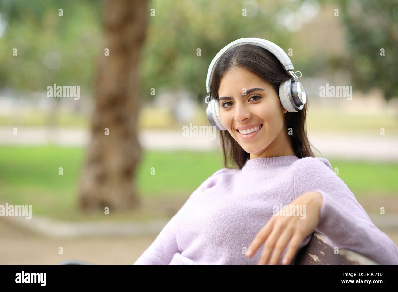 Glückliche Frau, die Musik hört und in die Kamera schaut, sitzt auf einer Bank in einem Park Stockfoto