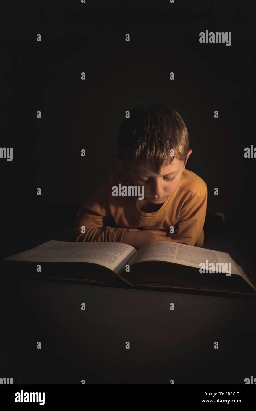 Das Kind liest ein Buch im Liegen, Dunkelheit um sich herum Stockfoto