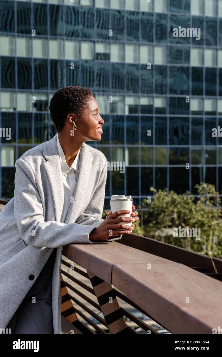 Vertikales Porträt einer attraktiven jungen afrikanischen Geschäftsfrau, die auf einem Balkongeländer liegt. Sie hält eine wiederverwendbare Kaffeetasse und hat Bürogebäude im Hintergrund Stockfoto