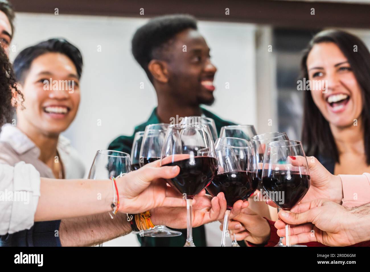Fröhliche multirassische Freunde, die Rotwein im Restaurant anstoßen. Eine Gruppe junger Leute, die Spaß am Indoor Winebar Tisch haben. Kulinarisches Leben und Freundschaft Stockfoto