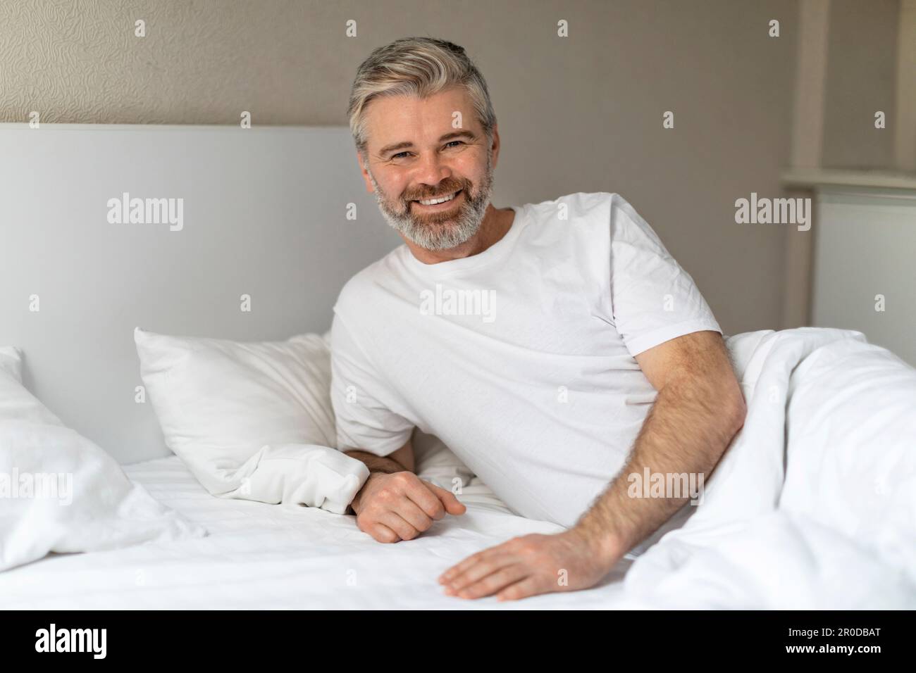 Glücklicher, gutaussehender Mann mittleren Alters, der im Bett sitzt und lächelt Stockfoto