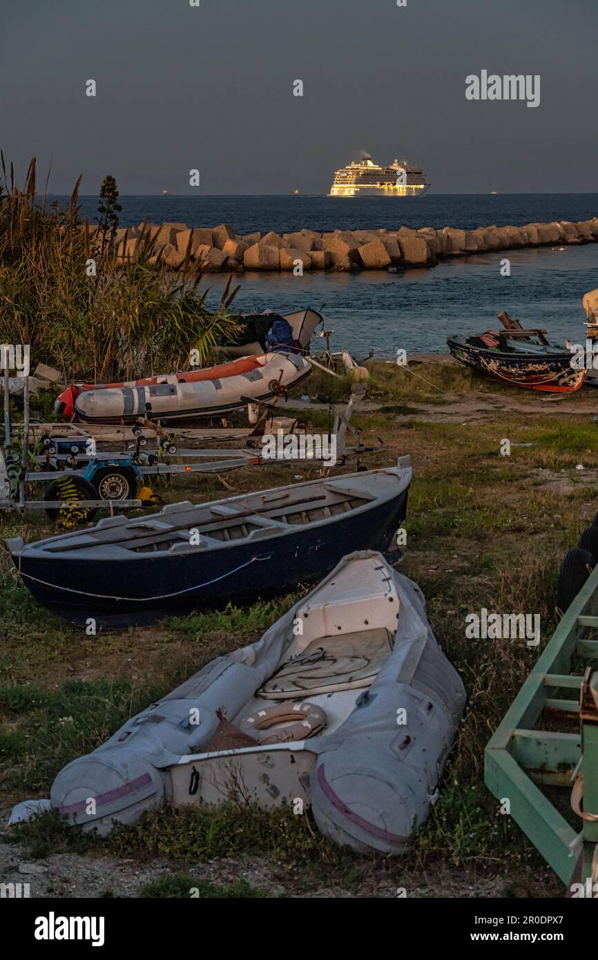 Das Kreuzfahrtschiff, beleuchtet durch das warme Licht des Sonnenuntergangs, überquert die Straße von Messina. Im Vordergrund zogen kleine Boote an Land. Kalabrien Stockfoto