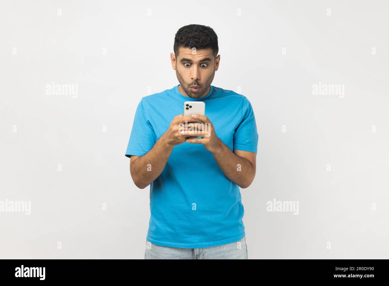 Portrait eines schockierten, unrasierten Mannes mit blauem T-Shirt, der mit dem Handy stand, soziale Netzwerke überprüfte, Nachrichten las. Studioaufnahmen im Innenbereich isoliert auf grauem Hintergrund. Stockfoto