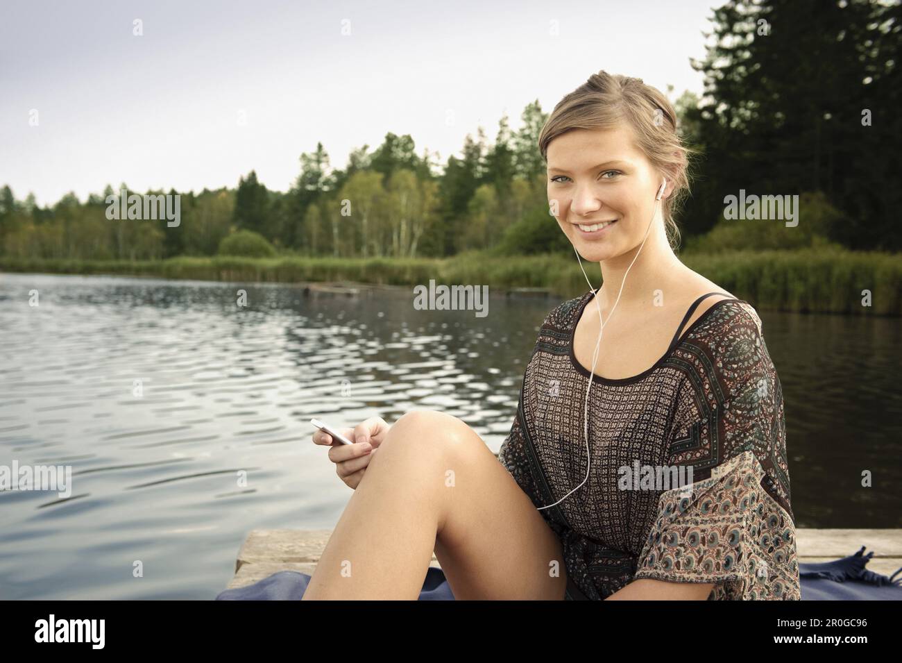 Junge Frau, die Musik hört, während sie auf einem Steg am Starnberger See in Bayern sitzt Stockfoto