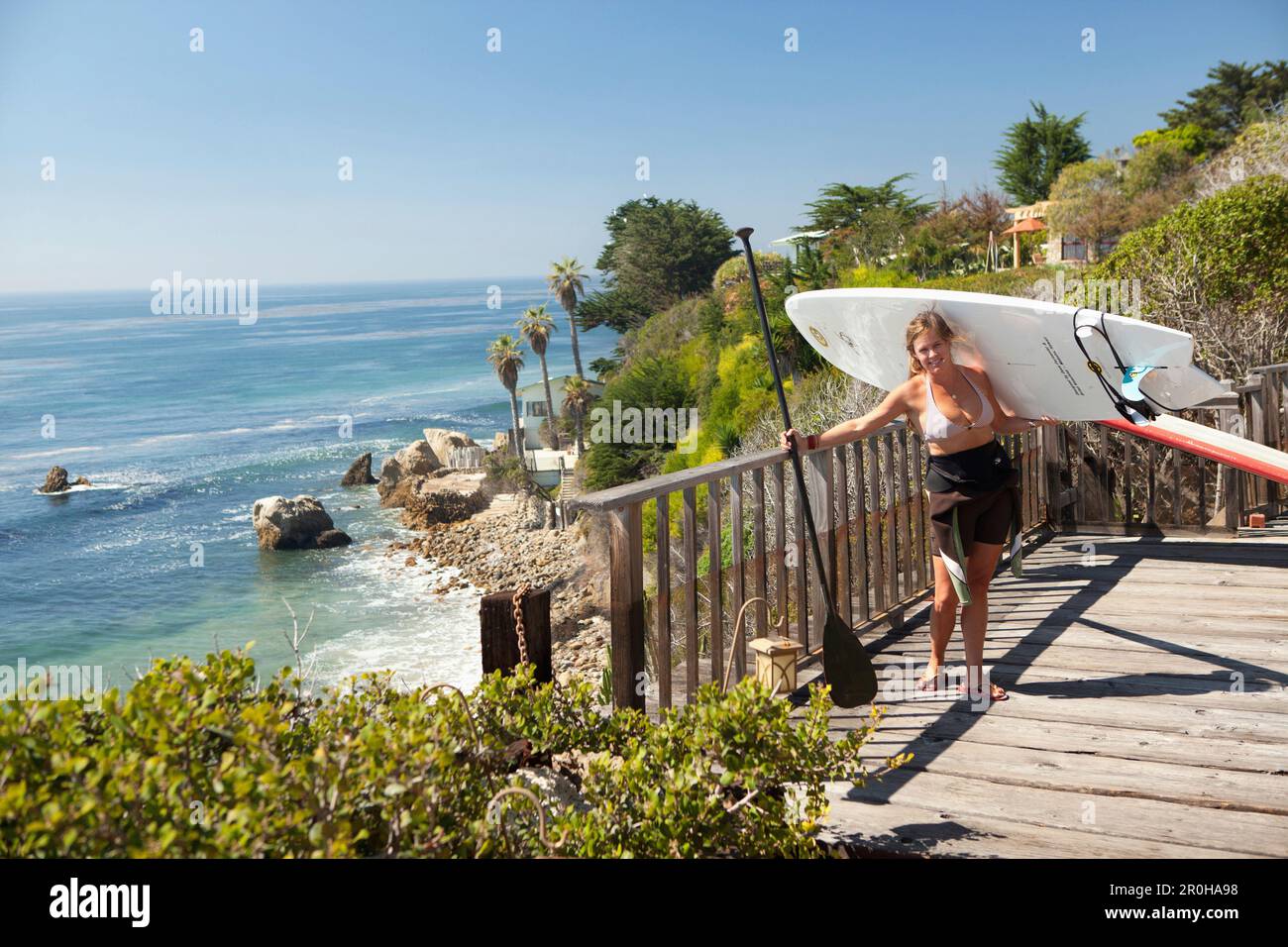 USA, Kalifornien, Malibu, El Pescador Beach – eine Sportlerin, die ihr Paddleboard in der Hand hält, macht sich bereit, zum Strand zu paddeln Stockfoto