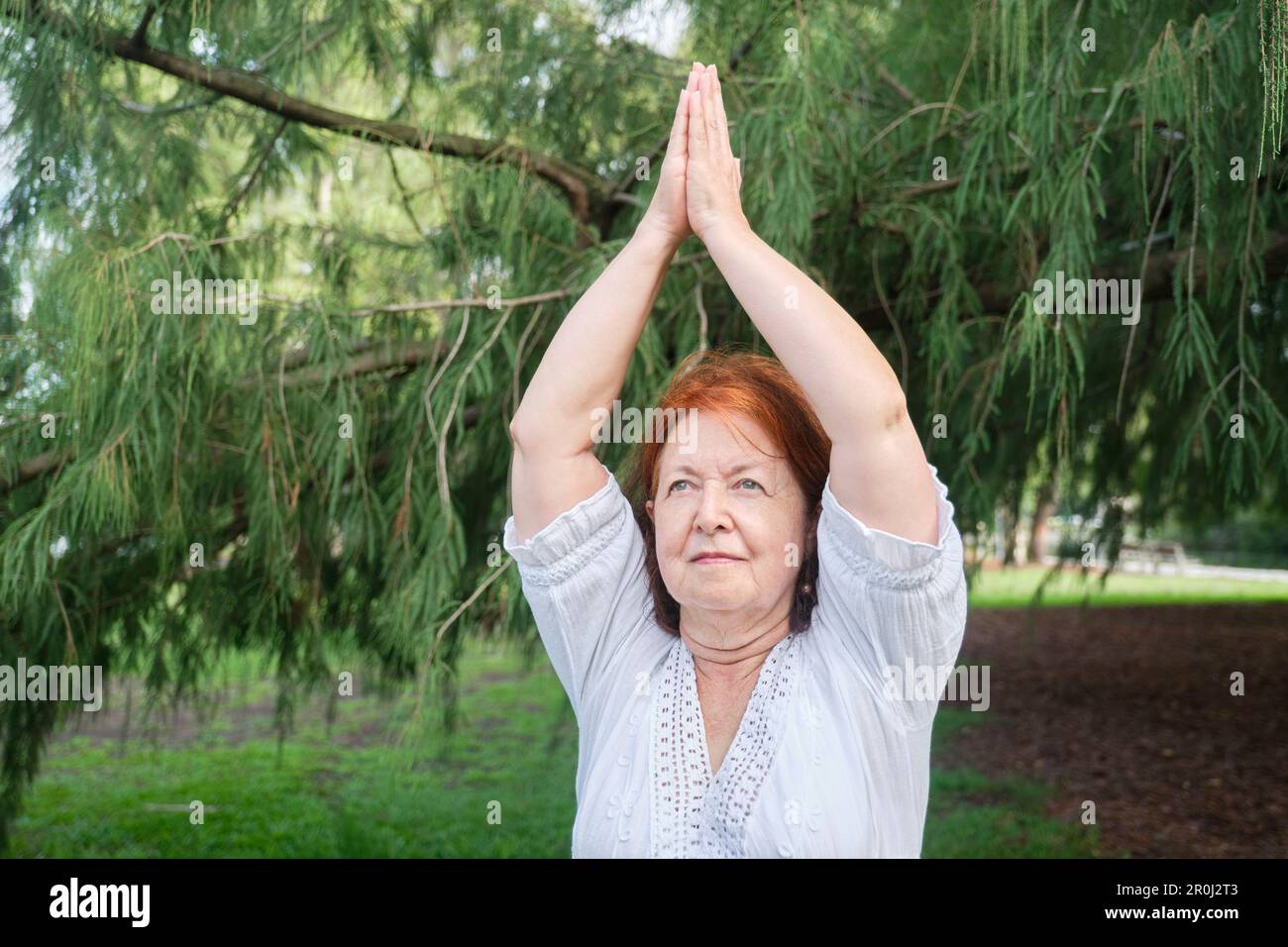 Ältere hispanische Frau in weißen Kleidern, die draußen Yoga in einem Park praktiziert. Vrksasana steht. Konzepte: Wellness, Vitalität, aktiv und gesund l Stockfoto