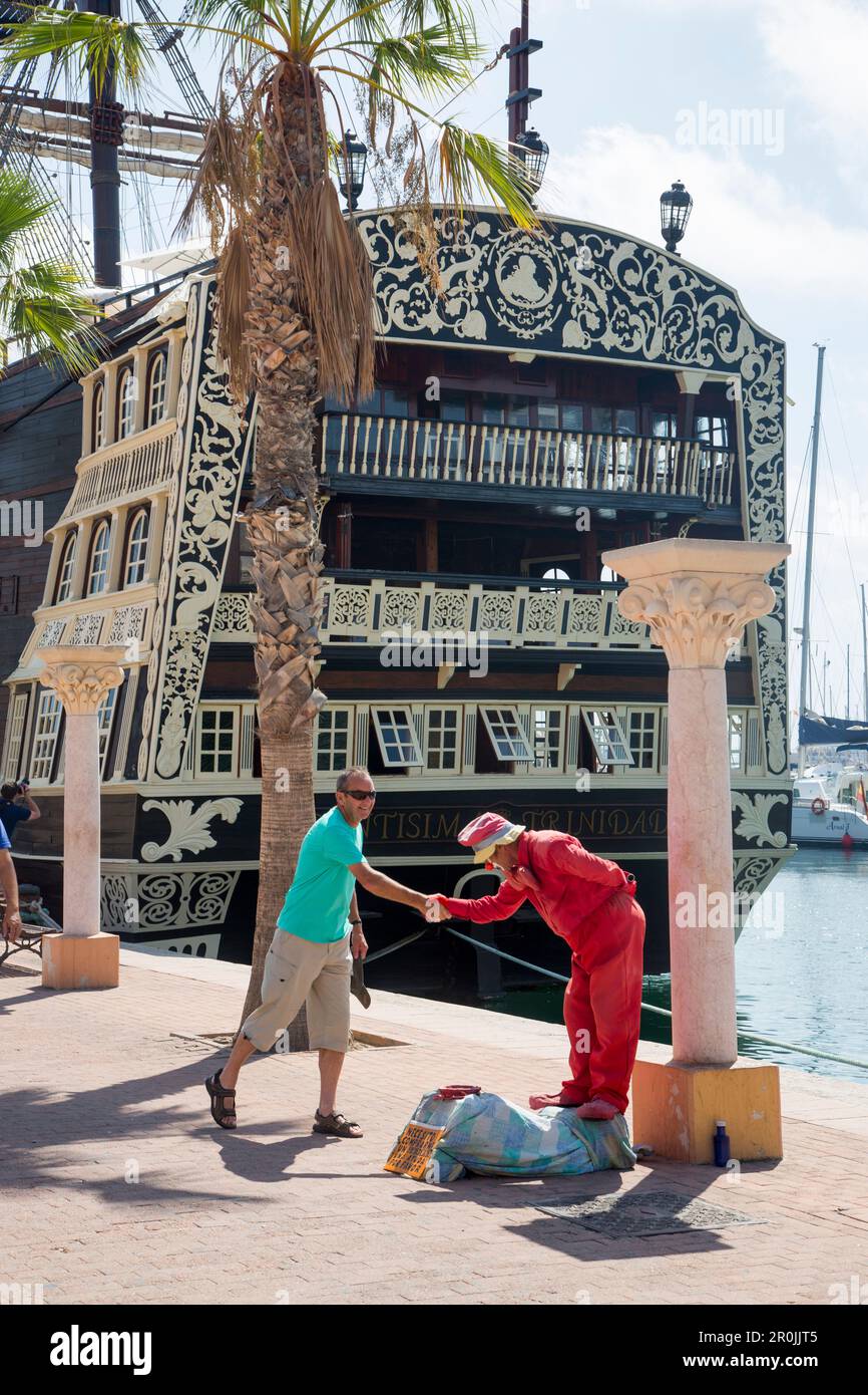 Der Clown Street Performer schüttelt die Hand mit dem Mann mit einer Nachbildung des historischen Segelschiffs Santisima Trinidad im Hafen dahinter, Alicante, Andalusien, Spanien Stockfoto