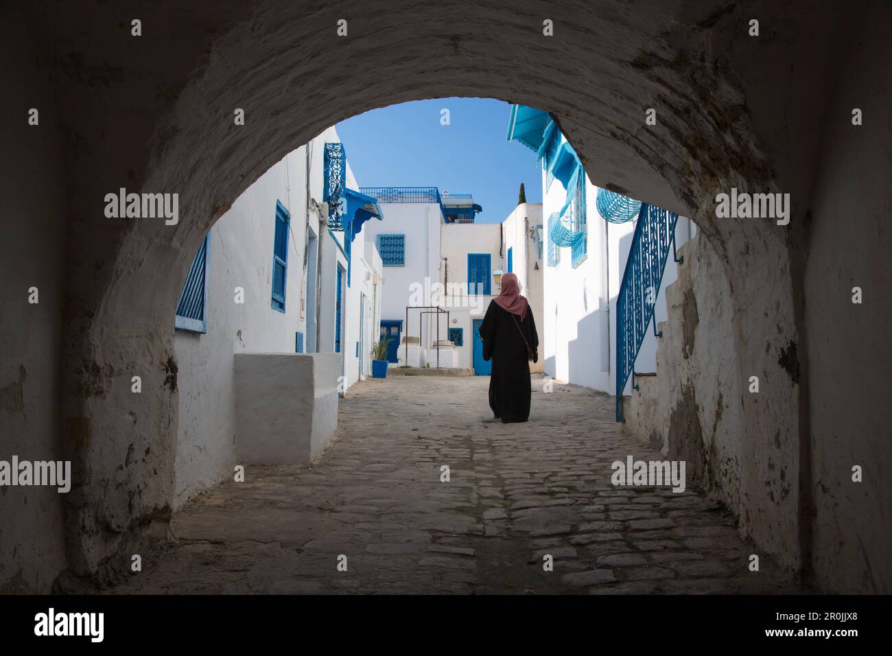 Arabierin geht durch einen Torbogen entlang eines Pfads von Gebäuden mit blauen Fenstern und weißen Wänden im Kunsthandwerksdorf Sidi Bou Said, Tunis, Tunesien Stockfoto