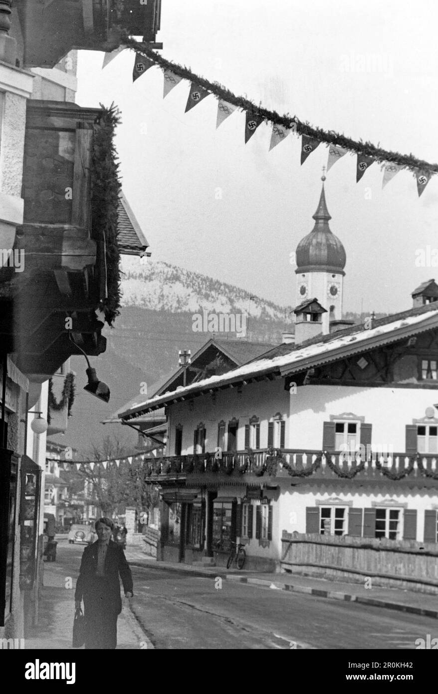 Eine Frau läuft die Straße entlang in Garmisch Partenkirchen, im Hintergrund die Kirche St Martin, über die Straße spannen sich Girlanden als Dekoration für die letzten Olympischen Winterspiele, deren Wimpel abwechselnd mit den Olympischen Ringen und dem Hakenkreuz der NSDAP versehen ist, 1936. Eine Frau geht die Straße hinunter in Garmisch Partenkirchen, im Hintergrund St. Martin's Church, Girlanden erstrecken sich über die Straße als Dekoration für die vergangenen Olympischen Winterspiele, ihre Wimpel wechseln zwischen den Olympischen Ringen und dem Hakenkreuz des NSDAP 1936 ab. Stockfoto