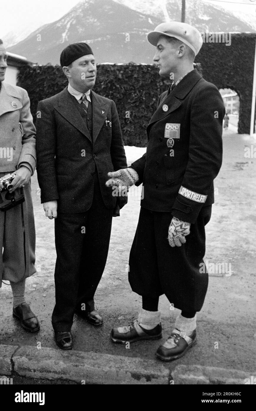 Ein Sporthelfer unterhält sich, an seine Jacke heftet das Reichssportabzeichen, ein Aufnäher mit den Olympischen Ringen und das Abzeichen der Olympischen Winterspiele in Garmisch Partenkirchen, 1936. Ein Sporthelfer ist im Gespräch, das Reich Sports Abzeichen, ein Patch mit den olympischen Ringen und das Abzeichen der Olympischen Winterspiele in Garmisch Partenkirchen sind an seiner Jacke, 1936, befestigt. Stockfoto