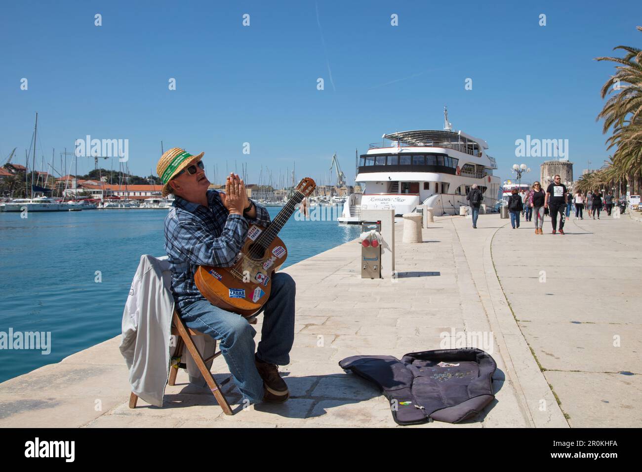 Der Straßenmusiker spielt Gitarre und tritt entlang der Uferpromenade auf. Dahinter befindet sich das Kreuzfahrtschiff MS Romantic Star (Reisebüro Mittelthurgau), Trogir, Split-D. Stockfoto