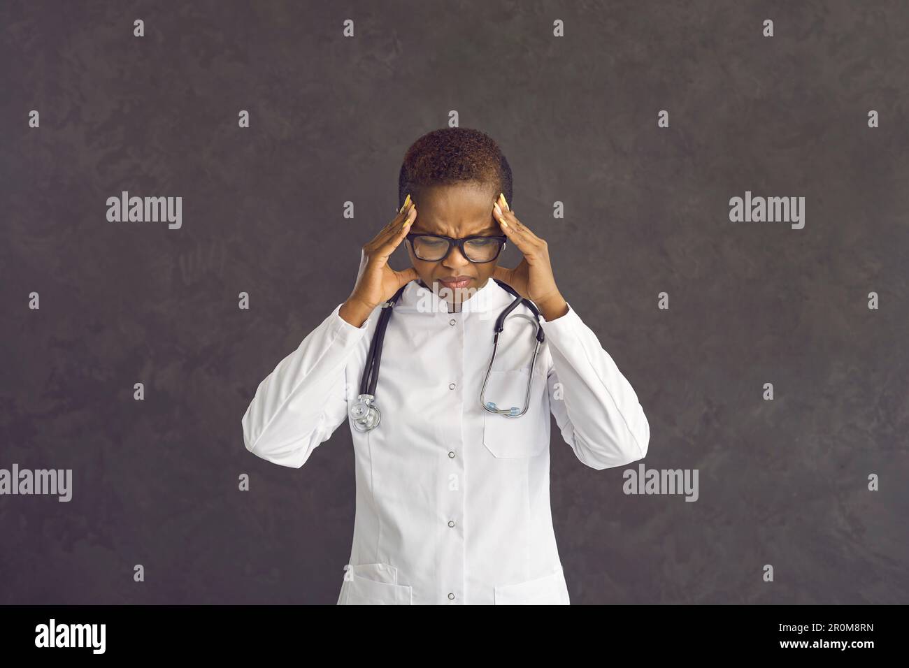 Erschöpfte, dunkelhäutige Ärztin steht auf einem Hintergrund mit grauer Betonwand. Stockfoto