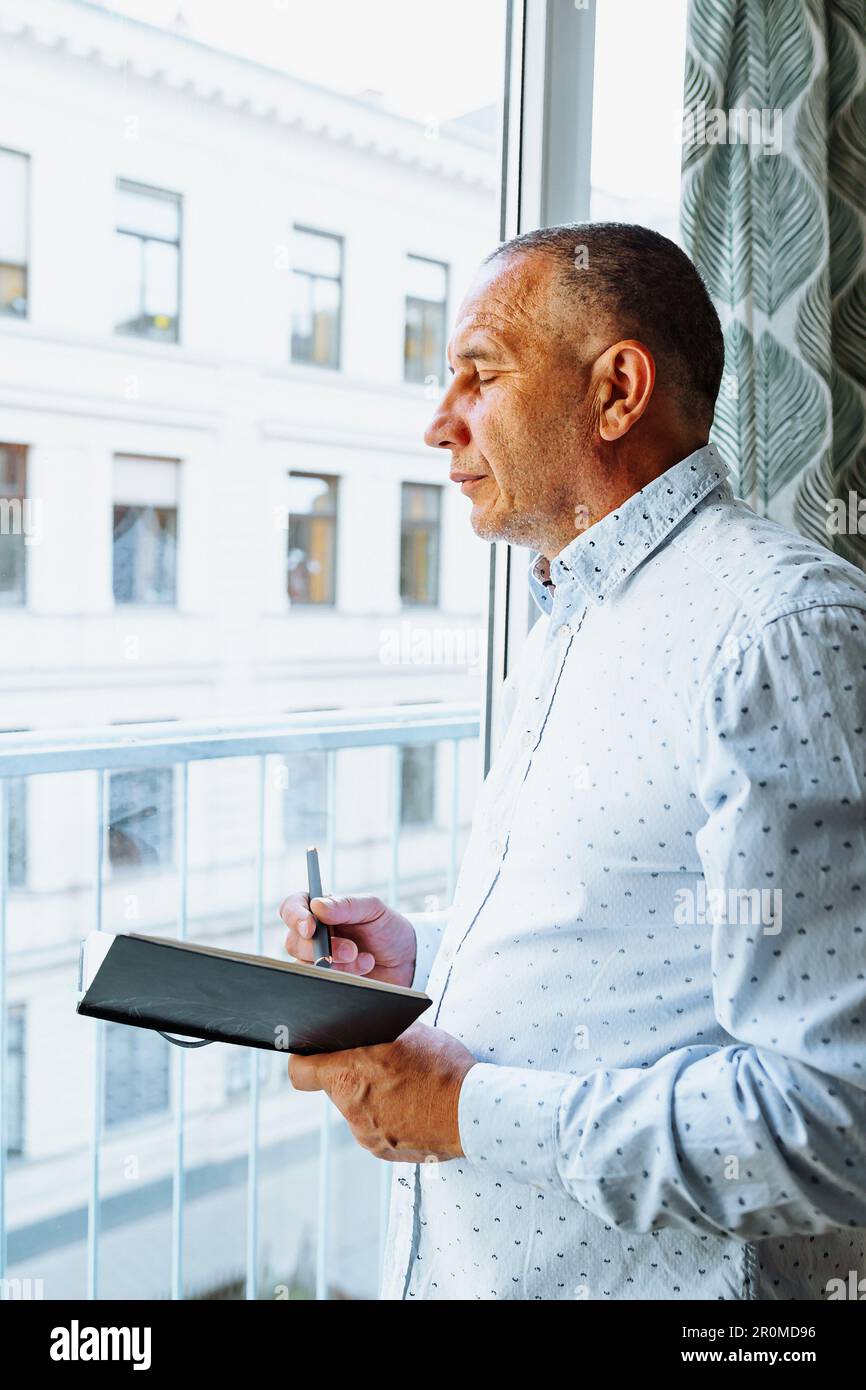 Attraktiver Mann im Hemd steht neben dem Fenster mit Notizblock Stockfoto
