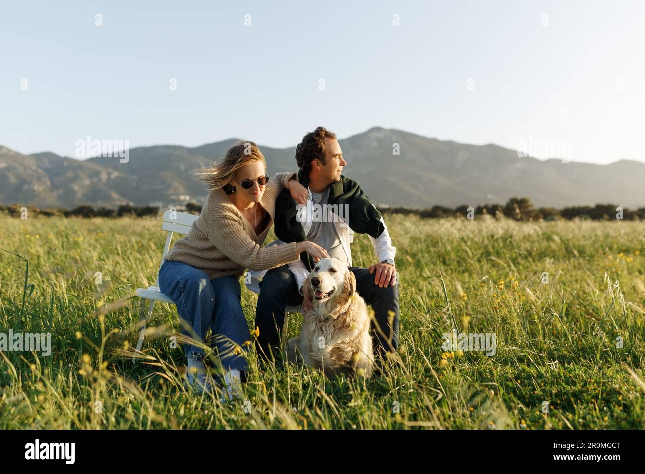 Ein junges glückliches Paar mit Hund, das auf Stühlen im Federrasenfeld am Sommertag bei Sonnenuntergang sitzt. Stockfoto