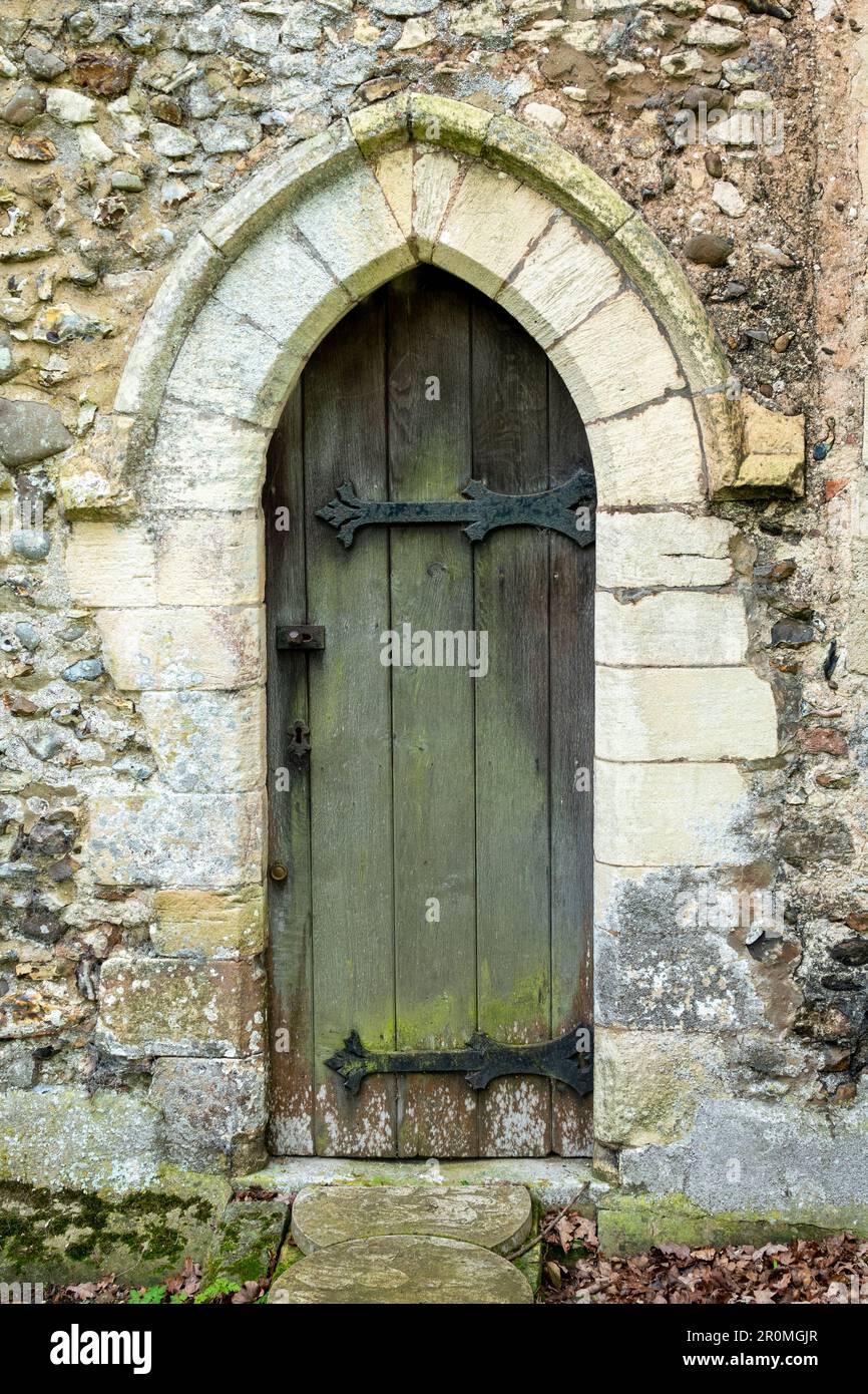 Kleine schmale Holzbogentür mit kunstvoll verzierten Scharnieren aus Gussmetall mit Steinumrandung in einer Steinmauer, St. Peter's Church Weston, Suffolk Stockfoto