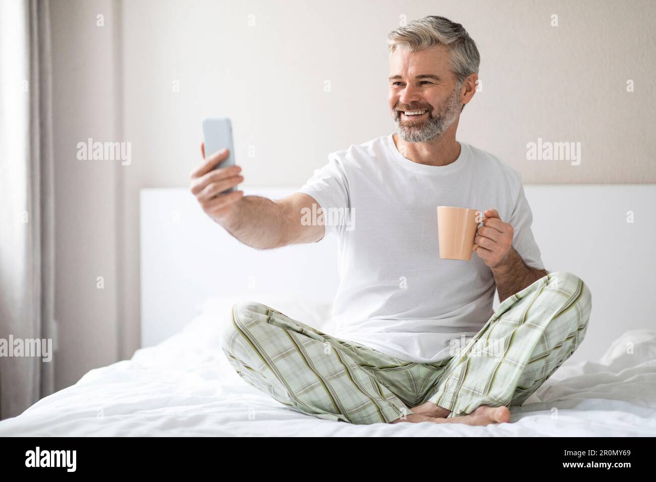 Glücklicher Mann, der im Bett telefoniert und Kaffee trinkt Stockfoto