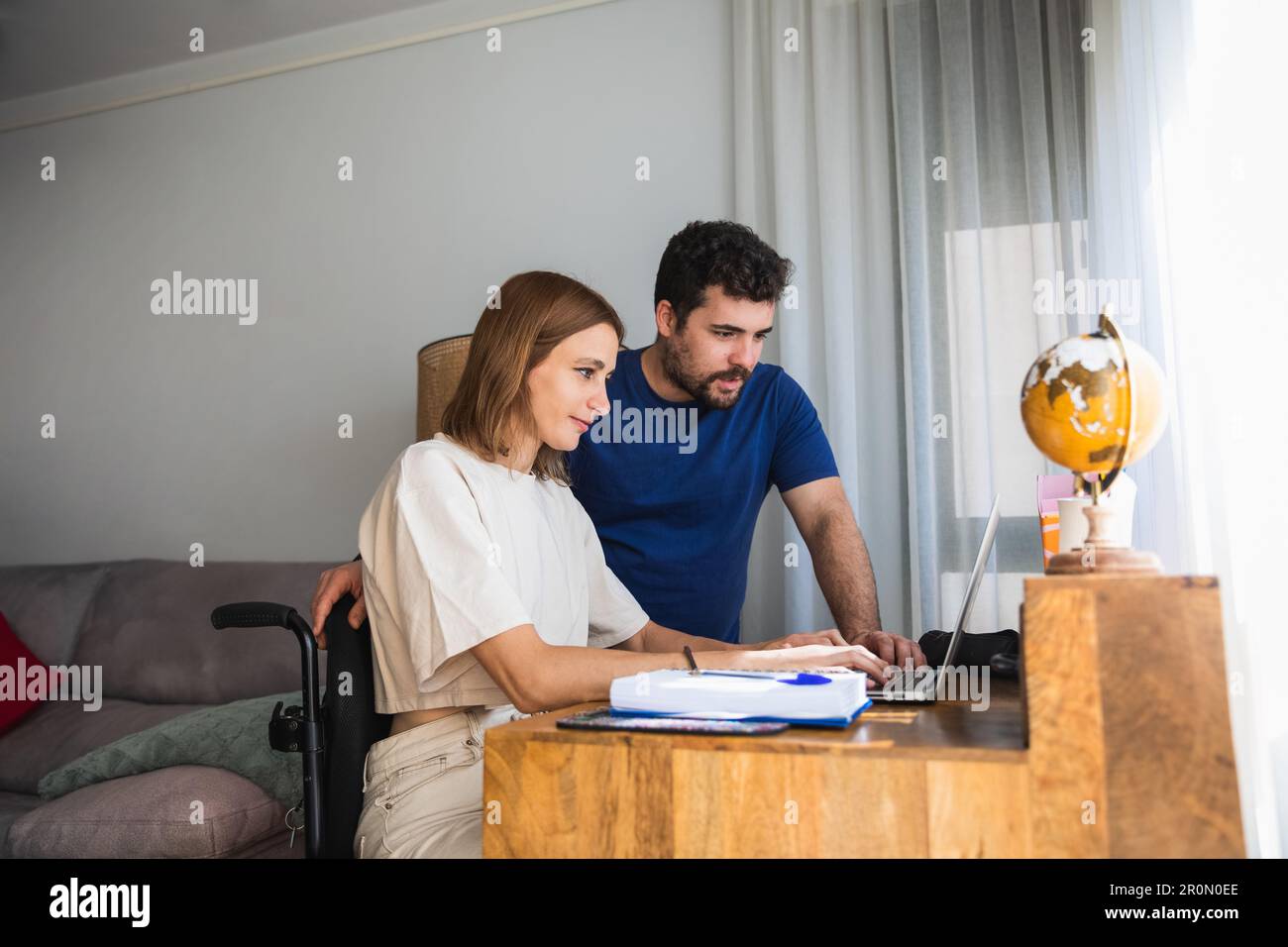 Liebenswerter, liebevoller Ehemann, der seiner Frau im Rollstuhl hilft, mit einem Büroprojekt zu Hause, das mit dem Laptop funktioniert Stockfoto