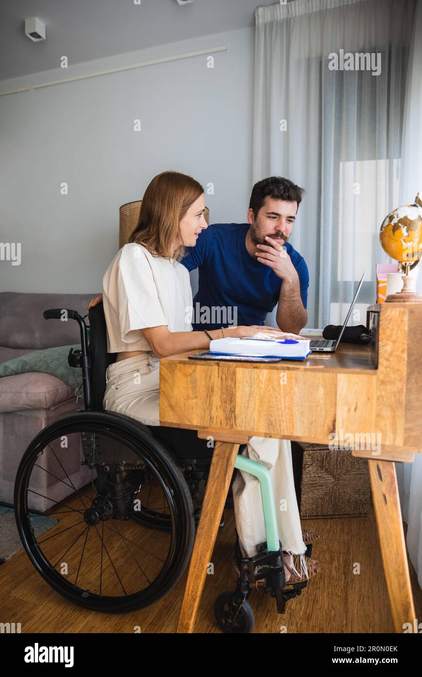 Liebenswerter, liebevoller Ehemann, der seiner Frau im Rollstuhl hilft, mit einem Büroprojekt zu Hause, das mit dem Laptop funktioniert Stockfoto
