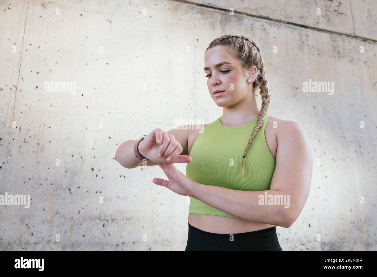 Beruhigen Sie junge Sportlerinnen in Sportbekleidung, die neben Betonwänden stehen und nach unten schauen, während sie beim Fitnesstraining eine moderne Smartwatch verwenden Stockfoto