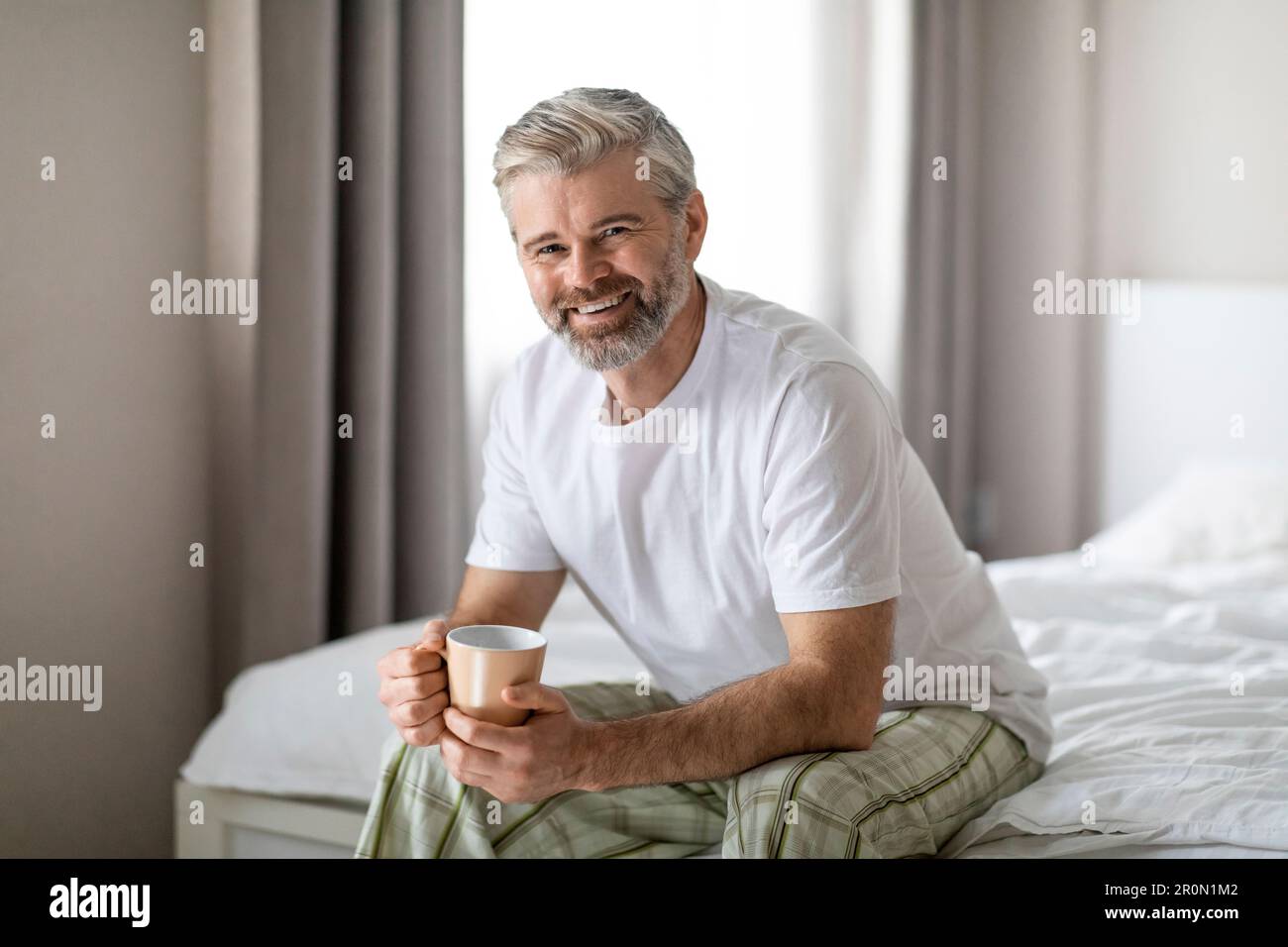 Glücklicher Mann mittleren Alters, der auf dem Bett sitzt und Kaffee trinkt Stockfoto