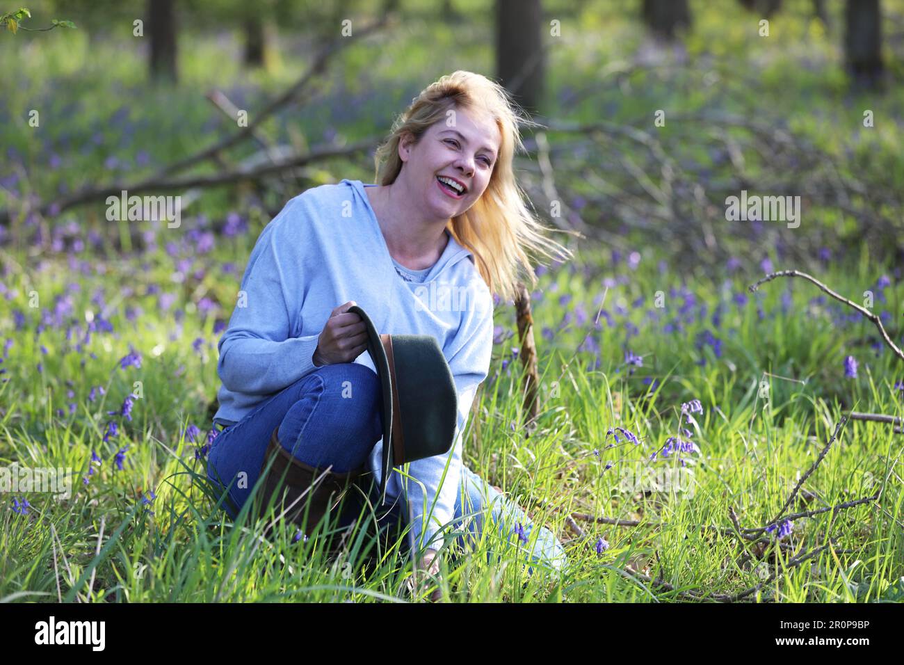 Eine Frau, die in einem Bluebell-Holz sitzt und einen Hut hält und lacht Stockfoto