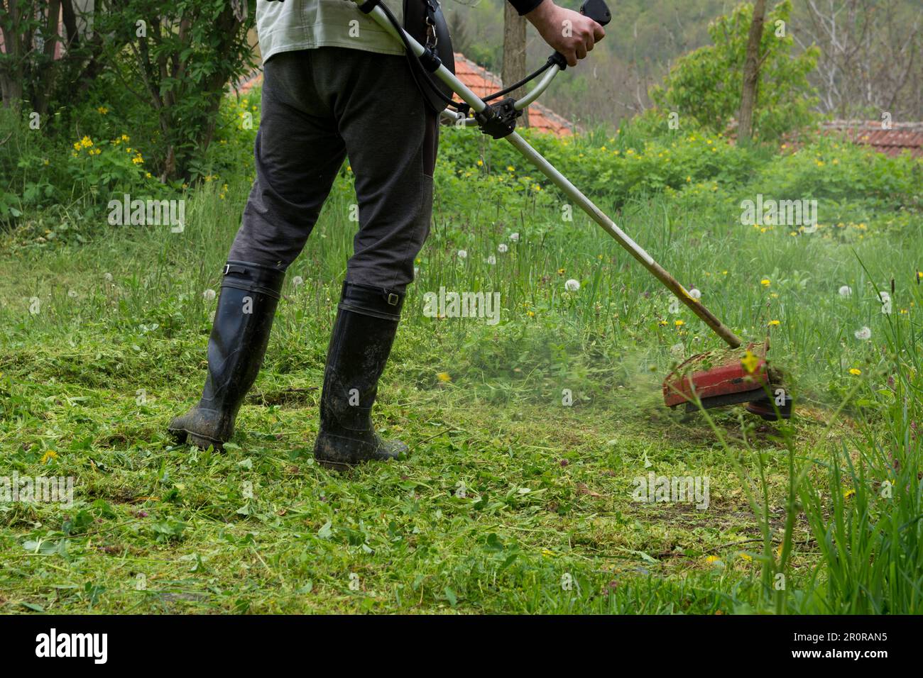 Ein Mann hält einen Rasentrimmer und schneidet an einem Frühlingsmorgen hohes Gras in seinem Garten. Nahaufnahme des Bauern in Stiefeln während der Arbeit auf der Wiese Stockfoto