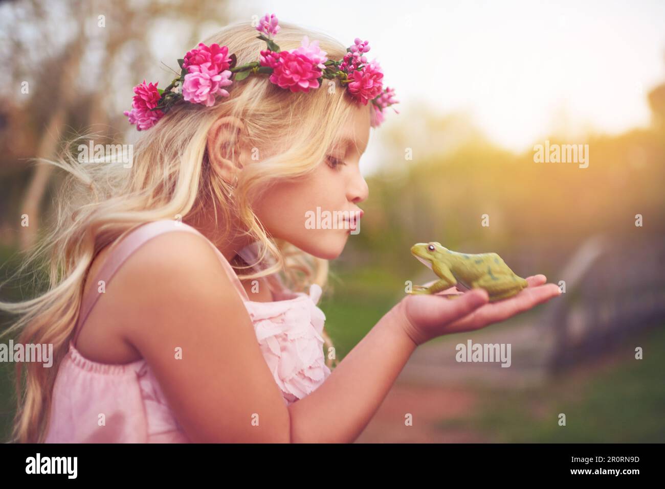Die Schönheit und das Biest. Ein fröhliches kleines Mädchen, das einen Frosch in der Hand hält und auf einen Kuss geht, während es draußen in der Natur steht. Stockfoto