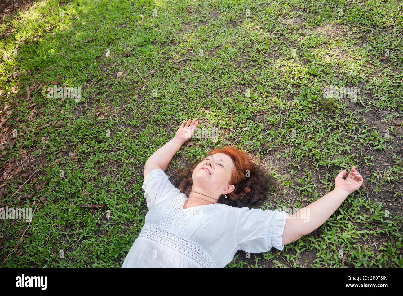 Reife hispanische Frau, die auf dem Gras liegt, mit ihren Armen in einer entspannten Position lächelt und einen Moment der Ruhe genießt. Stockfoto