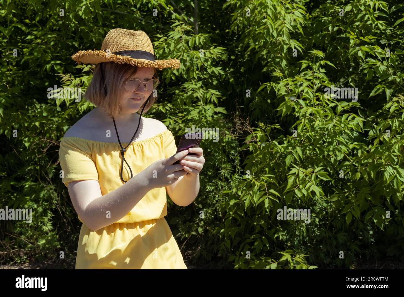 Ein asiatisches Mädchen mit Strohhut und gelbem Kleid, das in einem Park inmitten grüner Bäume eine Online-Nachricht auf ihrem Handy schreibt. Online-Shopping. Online-Schulung Stockfoto