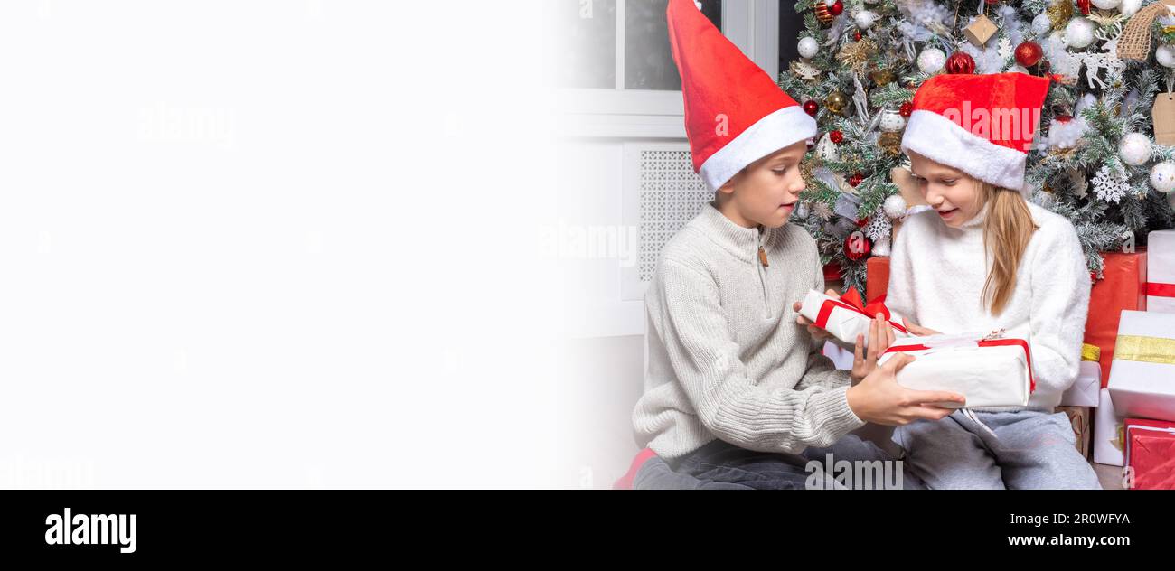 Süße, fröhliche, aufgeregte Kinder, Junge und Mädchen in weihnachtsmannhüten tauschen zu Hause Überraschungsgeschenke neben dem weihnachtsbaum aus. Schwester und Bruder schenken Weihnachtsgeschenke Stockfoto
