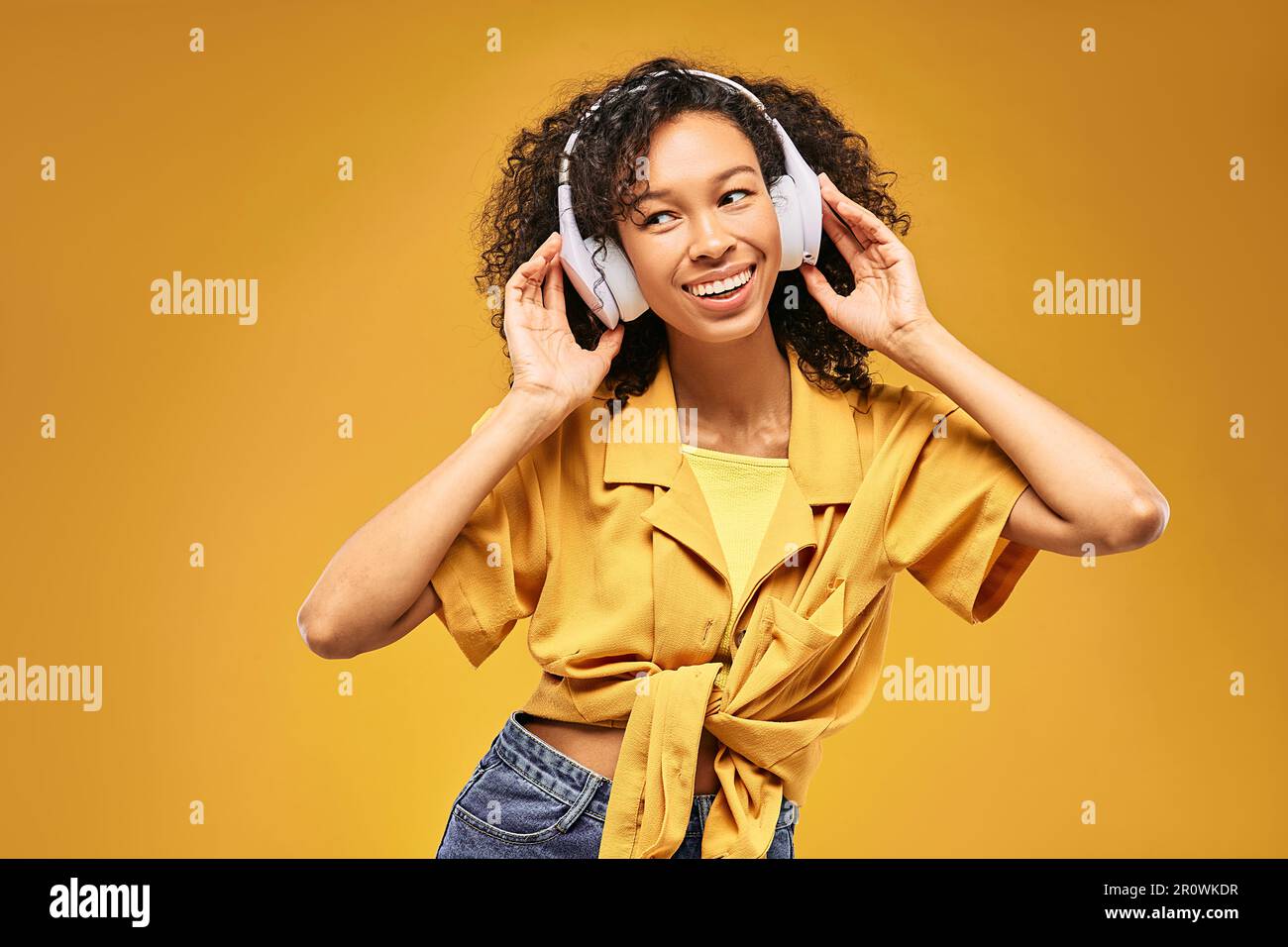 Glückliches afroamerikanisches Mädchen, das ihre Kopfhörer berührt, während es Musik hört, isoliert auf gelbem Hintergrund Stockfoto