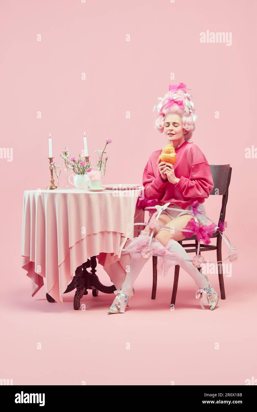 Porträt mit träumender Prinzessin, Königin mit großer Perücke, die anfängt, riesige Croissants auf rosa Hintergrund mit erstaunlichem Gesicht zu essen Stockfoto