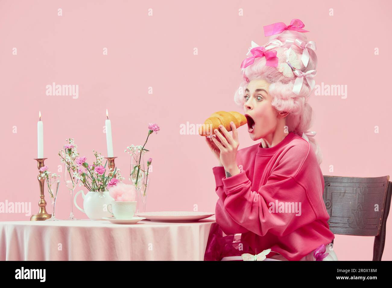 Porträt mit überraschter Prinzessin, Königin mit großer Perücke, die anfängt, riesige Croissants auf pinkfarbenem Hintergrund mit erstaunlichem Gesicht zu essen Stockfoto